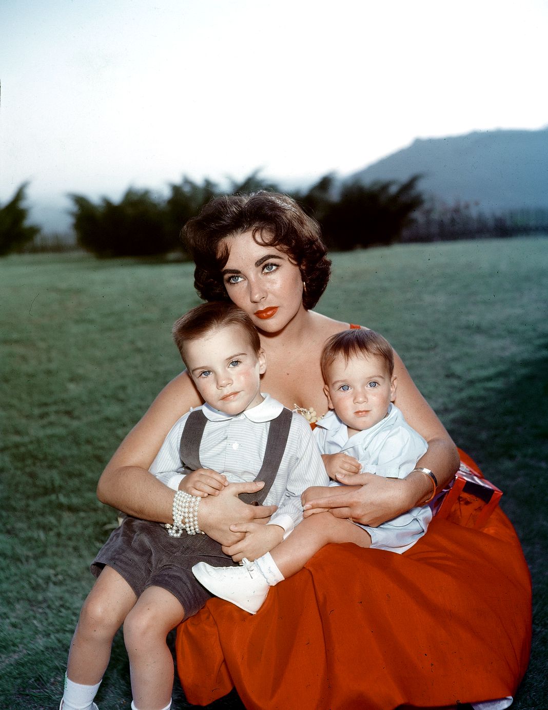 лизабет Тейлор сидит в красном вечернем платье со своими сыновьями Майклом и Кристофером Уайлдингом.