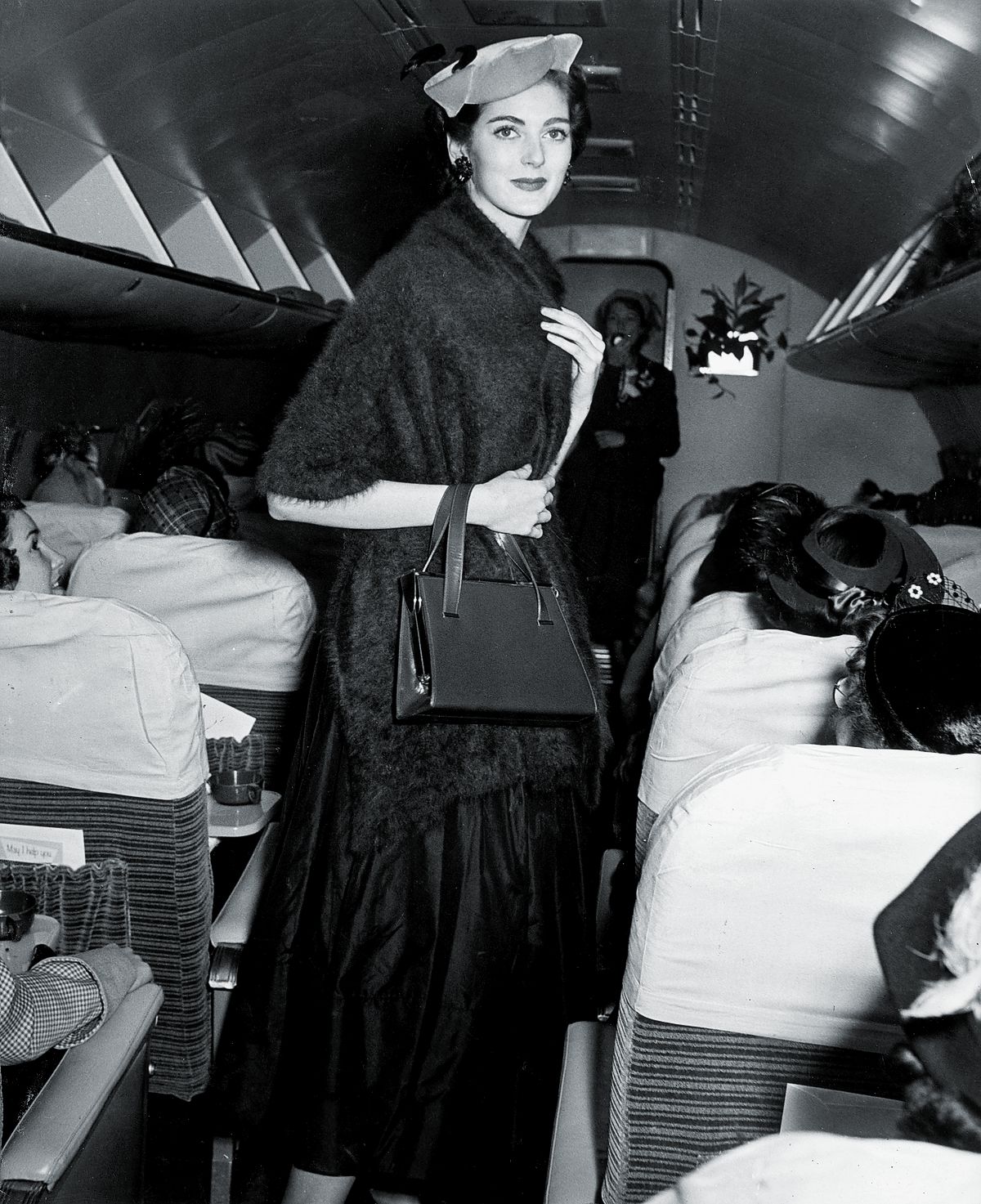 Кармен Делль’Орефиче в шляпе и мохеровом платке, а также с сумочкой идет по проходу самолета