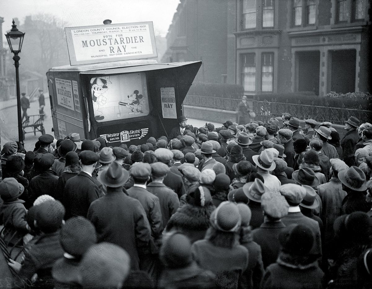 Во время избирательной кампании в Лондонский совет графства, Микки Маус отображается на маленьком экране на городской улице, надпись над экраном гласит: «Голосуйте за Мустардье Рэя»