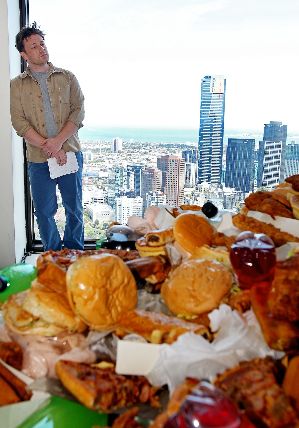 Джейми Оливер стоит перед столом, полным нездоровой пищи, в рамках проекта по борьбе с ожирением